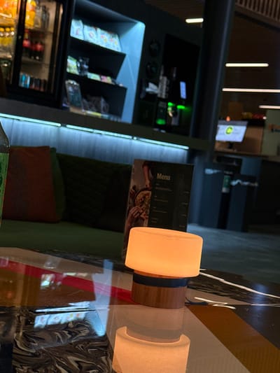 A blurred, slightly rotated image shows a table with a lamp, a menu, and a chair. The lamp is cylindrical with a tan shade and a brown base with a blue stripe. The menu has a dark cover with the word "Menu" printed on it. The chair appears to have green upholstery and a red cushion. In the background, there's a display case with drinks and snacks, illuminated by bright lights. A glimpse of a screen suggests a public setting.