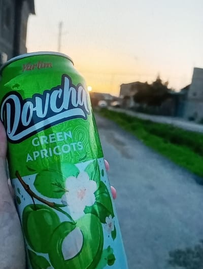 A left hand holds a green aluminum beverage can, prominently displaying "Dovcha" and "GREEN APRICOTS" on its label, along with illustrations of green apricots, a cross-section of an apricot, and white apricot blossoms. The background is out of focus, revealing an outdoor scene at either sunrise or sunset, indicated by a sky transitioning from light blue to orange and yellow. The sun's disc is visible near the horizon. A dark grey path or road extends into the distance on the right side of the frame, showing some wet patches. Blurred silhouettes of buildings and sparse vegetation are present on both sides of the background, along with a utility pole and a distant, faint light source. The location is Ortiugil, Uzbekistan. No other individuals are present.