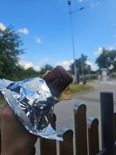 A person is holding a half-eaten chocolate snack bar, with its silver wrapper partially peeled back. The snack bar appears to have a biscuit base and a dark chocolate topping. The background is out of focus, showing a bright blue sky with scattered white clouds. Trees line the horizon, and a street light is visible in the distance. The scene suggests an outdoor setting, possibly a rest stop or roadside area, given the presence of what appears to be a road and some metal railings in the foreground. The lighting indicates it is daytime. The location is identified as Osikovitsa, Bulgaria.