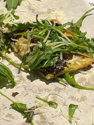 The image displays a close-up, top-down view of a single serving of food, likely an appetizer or small dish, resting on a light-colored, textured surface resembling parchment paper or a rustic plate. The main subject is a golden-brown, irregular piece of toasted bread or pastry, forming the base. This base is generously topped with a vibrant pile of fresh, green, leafy vegetables, which strongly appear to be arugula (rocket), with some leaves showing a slight sheen.
Scattered across the greens and the bread base are small, dark, irregular pieces, which could be chopped olives, dried fruit, or dark mushrooms. A fine layer of white, grated cheese, possibly Parmesan, is visible over the greens and other toppings. A few stray pieces of cheese and green leaves are also visible on the light surface surrounding the main food item.
The lighting in the image is bright and natural, casting soft shadows that enhance the three-dimensional texture of the food. There are no people visible, and no specific activity is taking place other than the presentation of the food. No visible text is present in the image. The scene is static and focuses solely on the appetizing qualities of the dish. The photograph was taken in Theewaterskloof Nu, South Africa.