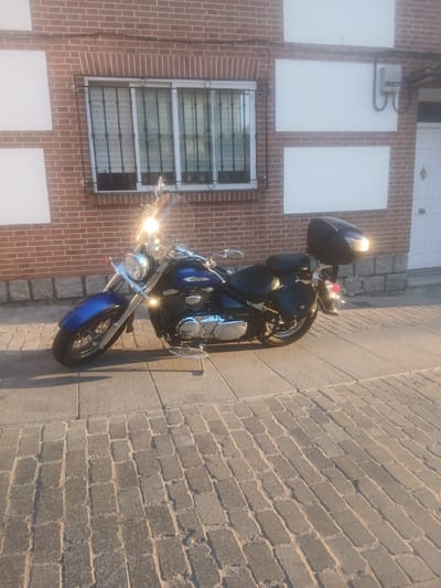 The image shows a blue and chrome motorcycle parked on a cobblestone sidewalk in front of a brick building. The motorcycle has a windshield, saddlebags, and a top case. The building has a window with metal bars and a white door. The scene appears to be in a city, possibly in Ávila, Spain, given the architectural style and cobblestone paving. The lighting suggests it is late afternoon or early evening. There are no people visible in the image.