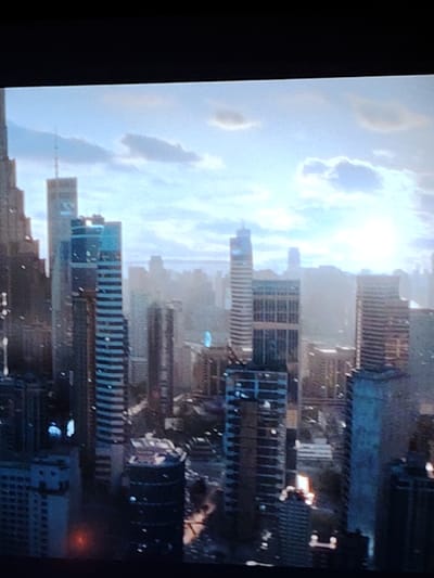 A skyline of tall buildings dominates the view. Many are rectangular and striped horizontally. Others are more streamlined with reflective windows. Clouds speckle the sky with the sun shining through. Mist appears between some buildings and in the distance. A few towers are topped with antenna. The light suggests it's either sunrise or sunset in a modern cityscape.