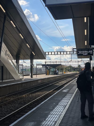 This is a vertically oriented, eye-level photograph taken at a train station platform. The scene depicts a modern train station with sleek, dark grey architecture, overhead power lines, and train tracks. The sky above is a vibrant blue with scattered white clouds, indicating fair weather.
In the foreground, the edge of a train platform is visible, characterized by a textured surface with raised circles. Two sets of train tracks run parallel to the platform. Overhead, a dark, angled structure with fluorescent lights runs along the top of the platform. The image is slightly tilted, giving a dynamic perspective.
On the platform, a person is seen from behind, wearing dark clothing and a backpack, looking towards the tracks. Further down the platform, other individuals are visible, some sitting and others standing. A digital display board shows train departure information, including destinations like "Innsbruck St. Pancras" and "London St. Pancras," along with times and platform numbers. The text on the display board reads: "let 1920 via innsion St.Panunes Expt 10.22 Caling at: coralich, ime H, Hasicube Park 2nd 18-32 sa Londun St. Pancras 18:2077". Another sign on a pillar indicates platform number "2". The presence of a sign reading "Inland" further suggests a train station setting. The overall atmosphere is that of a typical day at a busy transit hub.