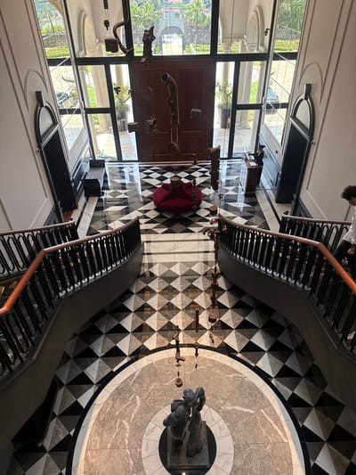 The image shows an overhead view of the interior of a building with striking architectural features. The floor is covered in a complex pattern of black and white diamonds surrounding a circular marble design. A staircase with black metal and wooden railings curves around this central pattern. In the foreground, on the left, is a large wooden door or panel with various metal pipes and gears attached. Large windows provide natural light, reflecting the outdoor scenery, which appears to be lush greenery possibly related to the landscape in Vila Verde. The room also contains modern furniture, including what appears to be a black table or console.
The architectural details and design elements suggest this is a high-end or architecturally significant building. The arrangement of objects and the elaborate floor design indicate careful planning and aesthetic consideration. No identifiable individuals are the focus of the image.