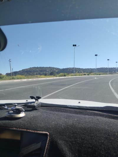 The image captures a view from inside a vehicle looking out through the windshield. The foreground shows a dashboard with a black fabric cover and a small silver helicopter-shaped air freshener. To the right of the air freshener is a black electronic device.
The road ahead is paved with asphalt and marked with white lines, indicating multiple lanes. On the left side of the road, there is a grassy embankment with a power line pole. In the distance, a gently sloping, tree-covered hill forms the horizon. Several modern streetlights with square fixtures are visible along the road. The sky is a clear, light blue.
The image does not provide any specific visual cues to pinpoint the location in the city of Myk, Uzbekistan.