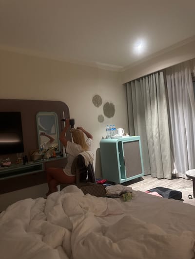 A hotel room interior in El Gouna, Egypt, features a blonde woman in a white t-shirt using a hair dryer in front of a mirror built into a light-colored, recessed cabinet. The cabinet also holds snacks and drinks. The room includes a bed with white linens, a luggage rack with a brown handbag, and a blue air purifier. The window is covered with horizontally-slatted, sheer curtains. The walls are light beige.