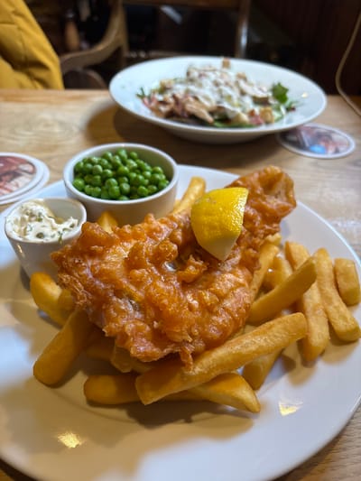 The image shows a plate of fish and chips. The fish is battered and fried, and a wedge of lemon sits on top. French fries are on the side of the fish. Additionally, there is a small bowl of peas and a small bowl of tartar sauce. To the left, there is also a small salad. The scene appears to be set on a wooden table, and the setting may be a pub or restaurant in the Vale of White Horse, United Kingdom.