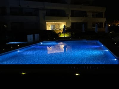 At night in the city of Playa Del Sol-villacana, Spain, an illuminated swimming pool glows with electric blue light. The pool is rectangular and tiled with small, dark blue squares. Lights are embedded in the pool's edge, casting a glow into the water. In the background, a multi-story building with balconies and windows is visible, partially illuminated by interior lights. Several closed white umbrellas are placed around the pool area, and the surrounding ground is dark and unlit, suggesting it's nighttime. A small jacuzzi or secondary pool is visible to the left of the main pool.