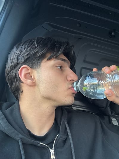 The image depicts a male individual inside a vehicle, located within the city of Kempten (Allgäu), Germany. The subject, a young adult male with dark, medium-length hair and light stubble on his face, is seen in profile from the right side, looking slightly upward and to the right. He is wearing a black hooded sweatshirt with a partially unzipped silver zipper over a black t-shirt. The male is actively drinking from a clear plastic water bottle held to his lips by his right hand. The bottle is partially filled with clear liquid, and the partial word "KRUM" is visible on its label. The background consists of dark, ribbed or paneled interior surfaces, characteristic of a vehicle's ceiling and upper wall, possibly a van or commercial vehicle.