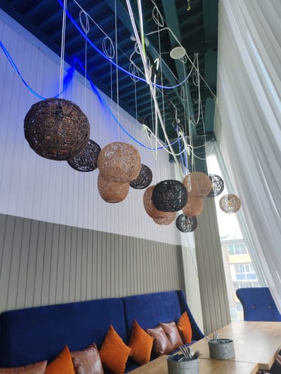 The image depicts the interior of a modern establishment, possibly a restaurant or cafe, featuring a long, plush blue sofa adorned with several orange and brown throw pillows. A light-colored wooden table is in front of the sofa, with two metal containers holding cutlery. Above, a series of woven spherical lamps hang from the ceiling, illuminated by ambient blue and white lighting. The walls are a combination of vertical gray paneling and white sections. A large, sheer white curtain drapes along the right side of the frame, partially obscuring a window that looks out onto a building with multiple floors and windows. The overall ambiance suggests a contemporary and stylish setting. The location is identified as Amursk, Russia.