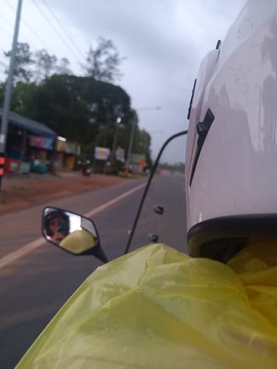 A person is riding a motorcycle on a road in Tambon Pak Phraek, Thailand. The rider is wearing a white helmet and a yellow raincoat. A person reflecting in the side mirror on the motorcycle is taking a photo of the rider with a smartphone. Buildings and trees are visible along the side of the road. The sky is overcast.