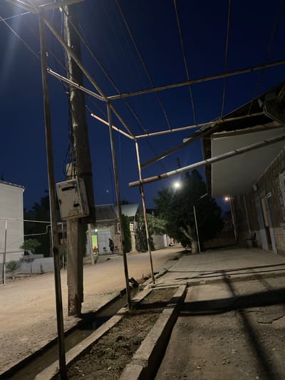 The image depicts a street scene at night in Verin Artashat, Armenia. A wooden utility pole stands on the left, supporting various wires and an old, weathered electrical box. To the right of the pole, a concrete drainage ditch runs along the edge of a paved sidewalk. In the background, a building with a green-lit interior and several chairs is visible, with a person standing near the doorway. Further down the street, more buildings and trees line the road, illuminated by streetlights. The overall environment suggests a quiet, residential area at dusk or night.