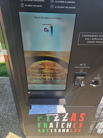 A black, self-service pizza vending machine is depicted outdoors in Fréjus, France. The machine features a large vertical digital display, a payment terminal, and product retrieval slots. The left side of the machine exhibits vertical green, white, and red stripes.

The digital display shows the procedural steps for ordering: "1) Je choisis mes produits" (I choose my products), "2) Je valide mon panier" (I validate my cart), "3) Je procède au paiement" (I proceed to payment), and "4) Je récupère mes produits" (I retrieve my products). The screen currently reads "Merci de votre commande" (Thank you for your order) alongside a graphic of a person with a pizza peel and thumbs-up icons. Below this, an image of a pizza cooking in a wood-fired oven is displayed with the text "Pizza artisanale précuite au feu de bois" (Artisanal pizza pre-cooked in a wood oven). "FLD tech" branding is visible at the screen's base.

To the right of the main display, a payment section instructs users to "Insérez la Carte Bancaire ou Payez sans contact" (Insert your bank card or Pay contactless). A physical payment terminal with a keypad, card slot, and contactless reader is installed. A slot for ticket retrieval is located immediately to its right. Above this section, text advertises ordering via the "SMART PIZZA" application.

At the bottom of the machine, a horizontal product retrieval slot, labeled "Machine sous surveillance" (Machine under surveillance) with a camera icon, is partially open, revealing a pizza box featuring line art of pizza slices and ingredients. The machine's lower front face is branded with large, multicolored text reading "PIZZAS FRAÎCHES ARTISANALES" (Fresh Artisanal Pizzas). The machine rests on a gravel or stone ground surface, with green foliage visible to the far left. No individuals are present.