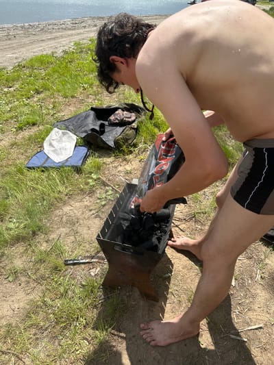 A person is outdoors preparing a charcoal grill. They are wearing dark, patterned swim trunks. The person is bent over the grill, holding a bag of charcoal and pouring it into the fire pit. The scene appears to be a grassy area near a body of water, with a sandy bank visible. The sun is shining, indicating it is daytime. Scattered around the grill are various items, including a black bag, a blue tarp, and a white cloth. The overall mood seems casual and relaxed, possibly a picnic or camping trip. The location context is provided as Pravda, Russia. There is some visible text on the charcoal bag, but it is partially obscured and difficult to read clearly. The weather appears to be sunny and warm.