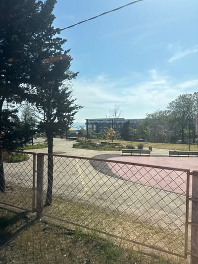 The image depicts an outdoor public space in Kholodnyi Rodnik, Russia, viewed through a wire mesh fence in the foreground. The fence, constructed of rusty chain-link material and supported by two concrete posts, separates a grassy area with sparse vegetation from a paved park or promenade beyond.
In the midground, a network of concrete paths with distinct pink-red and dark grey gravel sections forms a landscaped area. Multiple light-colored wooden benches with dark metal frames are positioned along these paths. Several trees of varying species, including dark evergreen and lighter deciduous types, are scattered throughout the park, casting shadows. A bright yellow, angular sculpture or play structure is visible to the left-center of the midground.
The background features a large, modern building with blue structural elements and numerous windows, suggesting a public or commercial facility. To the left of this building and extending towards the center, a expanse of blue water, likely the sea, is visible under a clear horizon. Additional dense green trees are present on the right side of the background.
The sky is bright blue with scattered thin white clouds, indicative of a clear, sunny day. A dark utility line or cable diagonally traverses the upper left portion of the sky. No individuals are present in the scene.