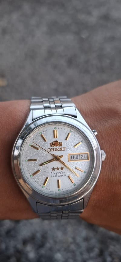 A close-up shot shows a man's wrist wearing a silver Orient watch. The watch has a white textured dial with gold hour markers and hands. A day and date window at the 3 o'clock position displays "THU 25". The watch band is also silver and made of segmented metal links. The background is out of focus and appears to be a grey, rough surface, possibly concrete or stone. The lighting suggests it is daytime, with a soft, diffused light. No specific emotions are conveyed, and the scene is focused on the watch. The visible text on the watch includes "ORIENT", "Crystal", "21JEWELS", and a small line of text at the bottom that is mostly illegible but appears to be manufacturing information.