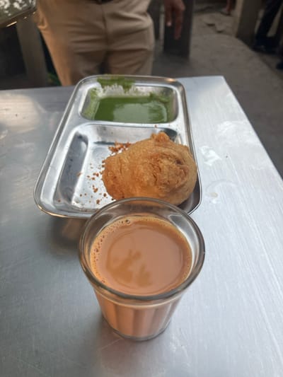 The image captures a serving of Indian street food and drink on a brushed metal table in Navi Mumbai, India. A clear glass, consistent with a "cutting chai" glass, holds a light brown, frothy beverage, identified as chai. Adjacent to the glass is a two-compartment stainless steel tray. One compartment contains a single, irregularly shaped, golden-brown fried item, likely a vada, surrounded by scattered crumbs. The adjacent compartment holds a quantity of vibrant green chutney. The metal table exhibits a brushed texture, minor scratches, and some small liquid splatters. In the blurred background, the lower body of an individual wearing light-colored trousers, possibly khaki, is visible. The immediate environment beyond the table appears to be an outdoor or semi-outdoor public space with an indistinct, uneven ground surface.