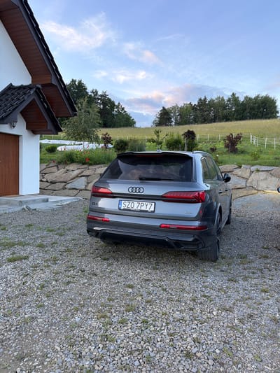 An Audi Q7 SUV is parked on a gravel driveway in front of a modern house with a dark tiled roof. The house has a white exterior and a wooden garage door. A stone wall, planted with some red flowers and greenery, separates the driveway from a grassy hill in the background. The hill is bordered by a white fence and dotted with trees. The sky is partly cloudy with hues of blue and pink. The license plate of the car reads "SZO 7PY7". The image appears to be taken in Limanowa, Poland, based on the license plate format and the style of the house and surrounding landscape.