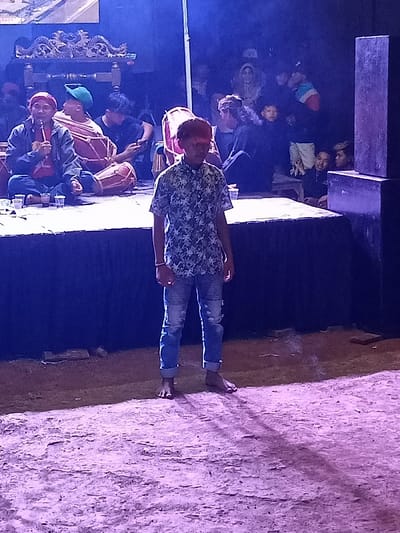 This image depicts a nighttime cultural event or performance in Kecamatan Cimaung, Indonesia. In the foreground, a young man, possibly a teenager, stands barefoot on a rough, unpaved surface, facing slightly to his left. He wears a short-sleeved, light-colored shirt with a dark, leafy pattern and ripped blue jeans with the cuffs rolled up. Dramatic purple and blue stage lighting illuminates him, casting a shadow behind him.

Behind the young man is a raised stage or platform where several individuals are seated. To the left, a man in a red head covering and blue jacket is holding a microphone, appearing to be singing or speaking. Next to him, another person wearing a green cap is playing a kendang, a traditional Indonesian drum. Other people are seated on the stage, some observing the performance, others seemingly engaged with phones or instruments. Traditional carved wooden elements, possibly part of a gamelan set, are visible on the stage, as are several clear plastic cups. A dark speaker box stands on the far right of the stage.

In the dimly lit background behind the stage, an audience is visible, mostly obscured by shadow, with some faces discernible as they watch the event. The artificial lighting creates a dynamic and somewhat mysterious atmosphere, characteristic of a live performance or cultural gathering at night. No visible text is present in the image.