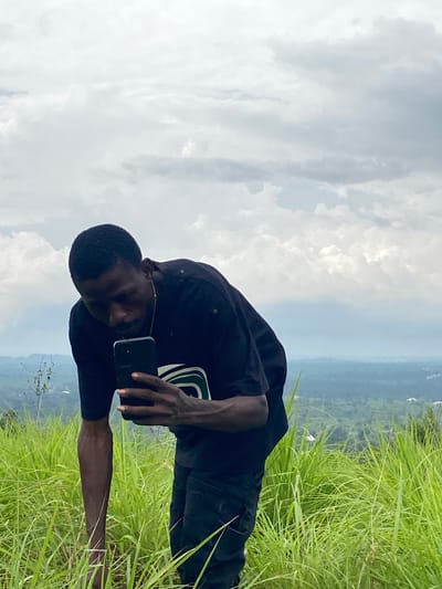 A young Black man is positioned in the foreground, standing in tall green grass on a hillside. He is wearing a black t-shirt and black pants, and is looking down at a black smartphone he is holding in his right hand. His left arm is bent, and his hand is resting on the grass. In the background, there is a lush, green landscape with rolling hills and a hazy sky filled with clouds. The setting appears to be outdoors in a rural or semi-rural area. It is daytime. The specific location is on a hillside in Nguzu Edda, Nigeria.