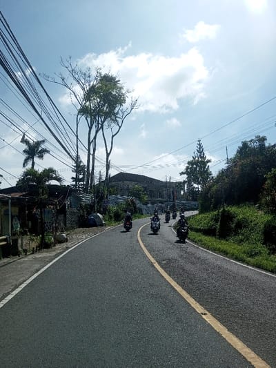The image presents a daytime scene on an asphalt road in Ban Phonhxavanh, Laos, curving gently to the right and appearing to ascend. The road features a solid white line marking the left shoulder and a continuous yellow line delineating the left side of the traffic lane. A procession of at least six motorcycles, predominantly scooters, is observed moving away from the viewer along the right side of the road's curve; each is operated by a helmeted individual. A seventh motorcycle is positioned further back in the left lane, also traveling away.

On the left side of the road, multiple utility poles support a dense network of black electrical and communication wires that crisscross the upper left quadrant of the frame. Below these lines, various structures are visible, including a small, low-rise building with a dark, corrugated roof in the foreground, adorned with colorful decorations and featuring a small green sign. A textured stone wall extends along the roadside, and a large, multi-story building, appearing to be under construction or in a state of disrepair with exposed concrete, is located further back. Interspersed among these structures are mature palm trees and several broadleaf trees, some exhibiting full green foliage while others display mostly bare branches.

The right side of the road is bordered by a sloping, densely vegetated embankment, covered with various green shrubs and foliage. A tall, slender coniferous tree stands prominently in the mid-ground on this side. The sky overhead is predominantly clear blue with scattered white cumulus clouds. Bright sunlight is evident, originating from the upper right portion of the frame, indicating midday conditions.
