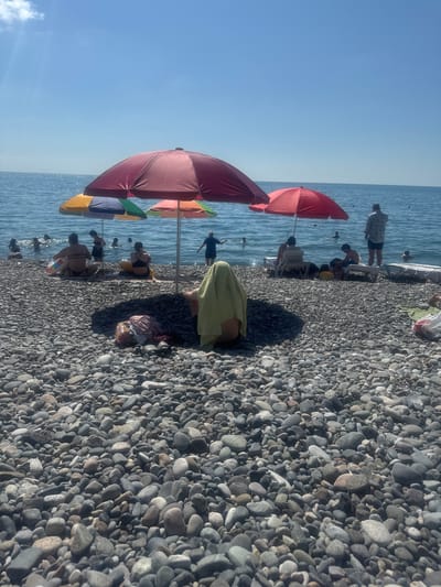 This media file shows a sunny day at a pebble beach in Sarpi, Georgia. The image is oriented vertically, with the sea on the left and the beach on the right. Several beach umbrellas, in shades of red, orange, green, blue, and yellow, are set up on the pebble shore, providing shade for beachgoers.
Numerous people are visible on the beach and in the shallow water. Some are sitting under the umbrellas, while others are wading in the sea. One person is partially covered by a yellow towel. Several individuals appear to be children, judging by their size. The water is calm and blue, reflecting the clear sky. The beach is covered in smooth, grey pebbles of various sizes. In the background, more people are scattered along the shoreline, and a few are swimming or playing in the water. The overall scene suggests a leisurely and relaxing day at the beach. The time of day appears to be daytime, given the bright sunlight and the shadows cast by the umbrellas. The weather seems to be warm and pleasant. There is no visible text in the image.