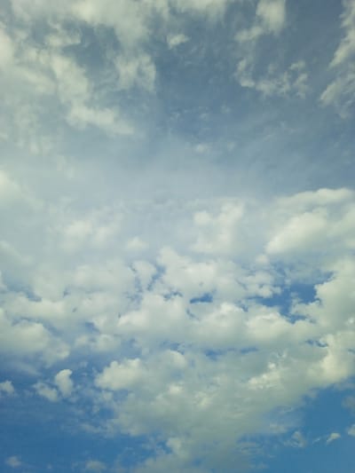 The image shows a vast expanse of sky filled with clouds. The sky is a gradient of blue, lighter towards the horizon and darker at the top, indicating it is daytime. There are two distinct layers of clouds. The lower layer consists of fluffy, cumulus clouds, white and bright, with distinct shapes and shadows, suggesting they are closer and more voluminous. Above these, a thinner, more diffused layer of cirrus or altostratus clouds is visible, appearing wispy and streaky, allowing more of the blue sky to show through. There are no visible landforms, structures, people, or animals in the image. The overall impression is one of an open sky, likely photographed from an outdoor location. There is no indication of the location in the city of Taran-Bazar, Kyrgyzstan.