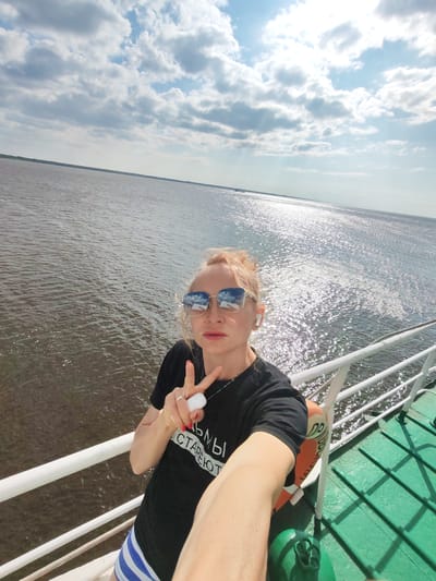 A woman with blonde hair tied back and wearing sunglasses and a black t-shirt is taking a selfie on a boat. She is making a peace sign with her right hand. The boat has a white railing and a green deck. In the background, there is a large body of water reflecting the sun, and a cloudy sky with patches of blue. The location is identified as Krokhino, Russia.