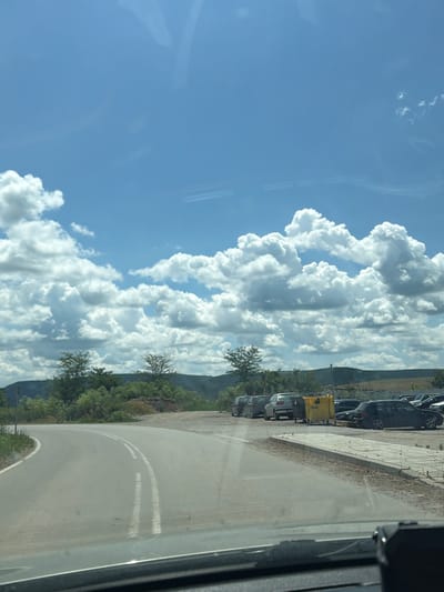 The media file shows a scenic view of a road winding through a lush green landscape under a bright blue sky dotted with large, fluffy white clouds. The scene appears to be captured from a moving vehicle, as indicated by the reflection on the glass in the foreground.

The road itself is paved with double white lines marking its lanes, and it curves gently to the right. To the left of the road, there is a paved walkway with square tiles, bordered by dry grass and dirt. Beyond the walkway, a line of parked cars is visible, including a yellow utility vehicle, several dark-colored cars, and a silver car. The cars are parked perpendicular to the road, suggesting a parking area.

The landscape is characterized by rolling hills covered in dense green vegetation and trees. The sky is a prominent feature, filled with dramatic, voluminous cumulus clouds that cast shadows, indicating a sunny day with variable cloud cover. The time of day appears to be daytime, likely late morning or early afternoon, given the bright sunlight and the clear visibility.

There is no visible text in the media file. The overall mood is one of serene natural beauty and a sense of journey or travel. The location context of Garchinovo, Bulgaria, suggests this could be a rural or suburban area in Eastern Europe.