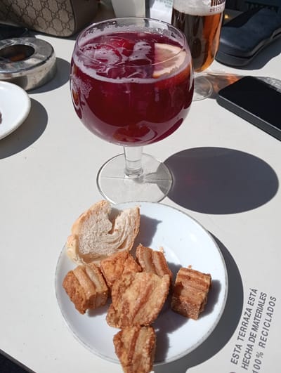 This image depicts a table scene on an outdoor terrace in El Espinar, Spain. The setting is illuminated by direct sunlight, producing sharp shadows on a light-colored table surface. In the foreground, a white ceramic plate holds six pieces of fried pork belly (torreznos) and a torn section of white bread. Centrally positioned, a large stemmed glass contains a red beverage, consistent with sangria, with ice cubes and a visible citrus slice (lemon or orange). To the left of the sangria glass, a metallic ashtray containing one cigarette butt is present. To the right, a glass of amber-colored beer with a head of foam is partially visible, alongside a black smartphone. A brown patterned bag is partially visible in the upper-left background. Text printed on the table surface reads: "ESTA TERRAZA ESTÁ HECHA DE MATERIALES 100% RECICLADOS," indicating the use of recycled materials for the terrace. No individuals are visible within the frame.