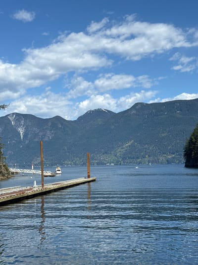 The image shows a scenic view of a body of water, likely a bay or inlet, with a mountainous landscape in the background. The sky is blue with scattered white clouds. A dock or pier extends into the water, with a boat or structure attached to it. The location is Bowen Island Municipality, Canada.