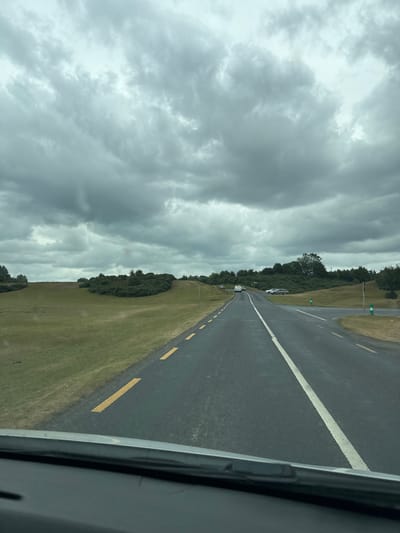 The image depicts a scene from within a vehicle on a cloudy day, looking out onto a rural road. The road is asphalt, with a white line dividing lanes and yellow dashes on the right side. To the left of the road, a grassy field slopes upwards, dotted with low-lying bushes and trees. In the distance, more trees and rolling hills are visible under a heavily overcast sky filled with dark, dramatic clouds. There are several cars parked or driving on the road, and some green marker posts are visible along the roadside. The overall impression is of a slightly damp, overcast day in a countryside setting, likely in Ireland given the reference to Curragh Edge.