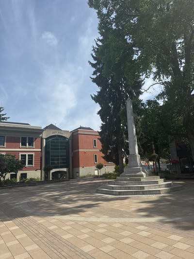 The media file captures a sunny outdoor public plaza in Port Coquitlam, Canada, featuring a prominent monument, a City Hall building, and extensive landscaping.

In the mid-ground, a tall, light-colored stone monument, resembling an obelisk with a pointed top, stands on a multi-tiered base. Inscriptions are visible on its facade, including the phrase "IN MEMORY OF" at the top of a panel, followed by several columns of names, suggesting it is a memorial. Lush green trees, including tall conifers and leafy deciduous trees, frame the monument and the left side of the scene, creating a natural backdrop.

To the right, a multi-story building with a distinctive architectural style serves as the City Hall. It features sections of red brick and grey concrete, with numerous windows. A large, curving glass facade marks the main entrance. The words "CITY HALL" are prominently displayed in large letters on the concrete section above this entrance.

The foreground and much of the mid-ground are covered by an expansive paved area made of light-colored, rectangular pavers arranged in a pattern. A series of curved concrete steps rise from this paved area towards the base of the monument. Shadows from the trees are cast across the paved ground, indicating bright sunshine. The sky above is light blue with scattered white clouds, suggesting clear and fair weather, likely during the mid-morning or early afternoon given the shadow angles. There are no people visible, and no specific event or activity is taking place.