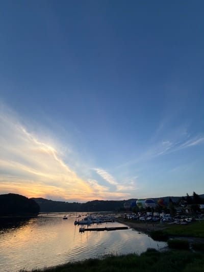 The image depicts a serene evening scene at the Gródek Lake, located in Gródek nad Dunajcem, Poland. The sky is a gradient of deep blue to light orange and yellow, indicating sunset or sunrise, with wispy clouds stretching across the expanse. The calm waters of the lake reflect the colors of the sky and the surrounding landscape.

On the left side of the image, a dark, tree-covered hill slopes down to the water. The lake is dotted with several sailboats and a few smaller vessels, some anchored near a wooden pier that extends into the water. The pier has a few small structures on it.

In the mid-ground and extending to the right, the shoreline is lined with a variety of hot air balloons of different colors and designs, some appearing to be in mid-inflation. Adjacent to the balloons is a parking area filled with numerous cars and recreational vehicles, suggesting an event or festival. Further back, a line of trees and buildings forms the backdrop to the scene. The ground in the foreground is covered with green grass and some darker vegetation. The overall atmosphere is tranquil and picturesque, capturing a moment of natural beauty and community gathering.