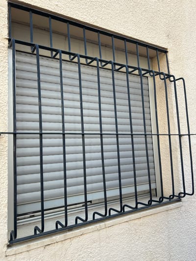 This media file captures a close-up, low-angle shot of a window with a black metal security grate. The window itself is covered by a closed, light-colored, possibly PVC or metal, roller blind with vertical ribbed detailing. The building's exterior wall, textured and painted in a light, creamy beige, surrounds the window frame. The metal grate is the most prominent object, featuring horizontal bars and decorative, zigzagging vertical elements on its left side. The grate appears to be made of thick metal rods, painted black. The lighting suggests it is daytime, with natural light illuminating the scene. There are no people or any discernible activity taking place. The setting appears to be an exterior wall of a building, likely residential, given the presence of a window and security grate. The location cues, such as the rough texture of the wall, are consistent with a Mediterranean or similar climate. No text is visible in the image.