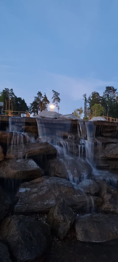 The image depicts a multi-tiered waterfall cascading down large rocks, with a clear blue sky in the background. Trees are visible on the left side of the image, suggesting a natural or landscaped outdoor setting. A single light fixture illuminates a portion of the trees and rocks, casting a soft glow. The time of day appears to be twilight or early evening, as indicated by the light sky and the illuminated light fixture. The water flow suggests movement and a gentle cascade. There are no visible people or text in the image. The overall mood is serene and tranquil. The provided location context places this scene in Shulgino, Russia.