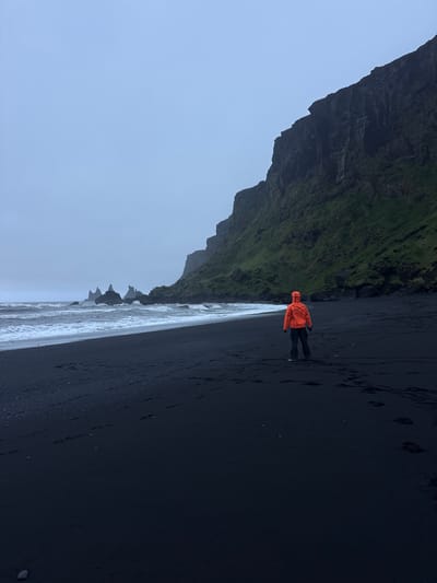 A wide-angle, tilted, medium shot depicts a person in an orange jacket standing on the black sand beach of Reynisfjara, near Vik, Iceland. The person faces away from the camera and towards the dark volcanic sand beach. The waves of the Atlantic Ocean roll in from the left side of the frame, adjacent to rock formations. To the left of the frame, a portion of the dark, moss-covered Reynisfjall mountain looms, meeting the sky. The weather appears overcast. Footprints mark the beach. The orientation of the image is rotated approximately 90 degrees counterclockwise.