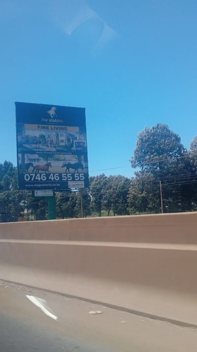A billboard advertises "The Stables" as "Fine Living," promoting a master-planned, gated community. It features images of upscale homes and horses. A telephone number, 0746 46 55 55, and the website address www.magnatecity.co.ke are prominently displayed. The billboard also includes the Magnate City logo, set against a backdrop of trees and a clear blue sky. The image is taken from a moving vehicle, focusing on the sign.