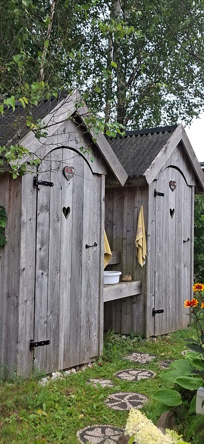 The image shows two wooden outhouses with corrugated roofs in Pihali, Estonia. The outhouses are made of weathered grey wood and have heart-shaped cutouts on their doors. One door is slightly ajar, revealing a yellow towel hanging inside and a white basin on a wooden shelf. The outhouses are situated in a grassy area with some stone path inserts leading away from them. A flowering plant with orange and yellow petals is visible in the foreground on the right side of the image. Trees with green leaves form a canopy above the outhouses.