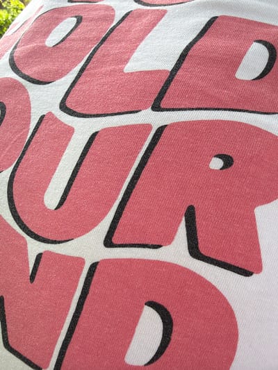 A close-up, slightly angled shot focuses on a white t-shirt with large, bold, pink letters outlined in black. The visible letters appear to spell out "SOLD OUR LOVE". The texture of the fabric is apparent, with visible knit lines. The background is softly blurred, suggesting an outdoor setting with green foliage, possibly trees or bushes, bathed in natural light. The image does not contain any elements that would suggest a specific location in Poroshkino, Russia.