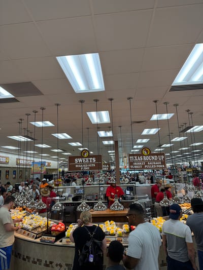 The image depicts the interior of a Bucee's convenience store located in Florence, South Carolina, United States. The scene captures a busy food service area with customers and employees. Several signs are visible, indicating food offerings such as "SWEETS SUGARIED NUTS FUDGE" and "TEXAS ROUND UP BRISKET SAUSAGE TURKEY PULLED PORK." Customers are standing in line, browsing food displays, and interacting with employees who are behind the counter preparing and serving food. The environment appears to be well-lit with fluorescent lighting from the ceiling. The food display features various wrapped items, possibly sandwiches or snacks, along with some bags of chips. The overall atmosphere suggests a retail and food service environment common in large convenience stores.