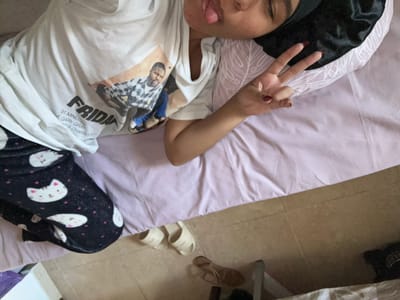 The image shows a person lying on a bed, wearing pajamas with a cat print and a t-shirt with a graphic of Ice Cube from the movie "Friday". The person is sticking their tongue out and making a peace sign with their right hand. The bed is covered with a light purple sheet. Beside the bed, on the tiled floor, are a pair of white slippers and a pair of sandals. The overall scene is casual and intimate.

The location is identified as Pueblo Salinas, Spain. This is a small village in the province of Almería, Andalusia, known for its agricultural production, particularly olives and vegetables. The environment depicted in the image, with tiled floors and simple furnishings, is consistent with a residential interior in a Spanish village. The lighting appears to be natural, coming from an unseen window or overhead source.