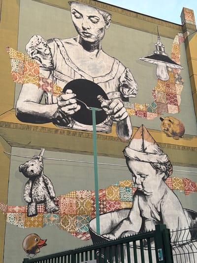 The image shows a mural on a building in Aubervilliers, France. The mural features a collage-like design with grayscale drawings of a figure pouring liquid into a dark circle, disembodied hands, a toy bear hanging by a string, and a lightbulb resembling a syringe. A yellow piggy bank is floating above the figures. The background of the mural is a patchwork of patterned tiles and vertical lines. The edge of a fire escape is visible on the right side of the image.