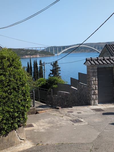 The image shows a view from a paved pathway overlooking a body of water with a large arched bridge in the distance. A lush green bush occupies the left foreground, with a stone and metal fence lining a pathway that descends into the foreground. A dark gray gate is visible on the right, constructed with stone pillars. Overhead power lines are visible against the clear blue sky. The environment suggests a sunny day in a coastal area, consistent with Kraljevica, Croatia, known for its bridges spanning across bays. No people are visible in the image.