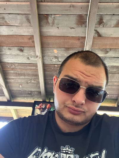 The image is a close-up selfie of a man wearing sunglasses and a black t-shirt. He is indoors, under a wooden ceiling. The man has short, dark hair and a light beard. He is looking towards the camera with a neutral expression. The background consists of wooden planks of the ceiling. A portion of a television screen is visible in the lower left of the image, displaying a picture of two men. The location is identified as Dragoevo, Bulgaria.