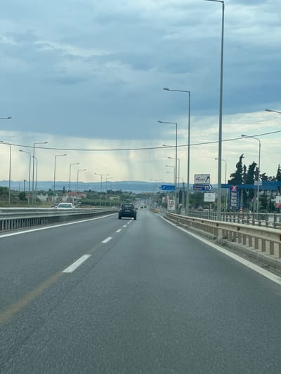 The image depicts a highway scene in Nea Moudania, Greece. The photograph is taken from a vehicle moving along the right lane of the highway. The road is grey asphalt marked with white lines. A dark-colored car is visible ahead in the same lane. To the left of the highway, there are multiple streetlights lining the road. Further beyond, there is a landscape with greenery, buildings, and hills in the distance. The sky is partly cloudy with a light blue hue. Gas stations and road signs are visible at a distance.