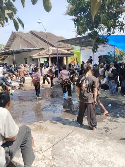 The image depicts an outdoor scene in Botok, Indonesia, likely a community gathering or performance. Several individuals, some dressed in traditional attire featuring striped shirts and dark pants, are present. Two individuals in the foreground appear to be in mid-performance, with one holding a prop or costume. A crowd of onlookers stands in the background, observing the activity. Buildings with tiled roofs are visible, suggesting a residential or communal area. The ground is wet, possibly from recent rain or cleaning, and scattered debris is present. A banner with Indonesian text is displayed in the background, indicating a special event. The sky is clear and blue, suggesting fair weather.