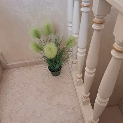 The image depicts an indoor scene, possibly a stairwell or hallway, with a focus on decorative elements. A potted plant with long, green, grass-like foliage and fluffy, spherical light green seed heads sits on a marble-patterned floor. To the right, a white wooden banister with gold accents lines the edge of the scene. The wall behind the plant has a textured, neutral finish. The perspective suggests the photo was taken from an elevated position, looking downwards at the plant and banister. The location is identified as Tairove, Ukraine. There are no people or animals visible in the image.