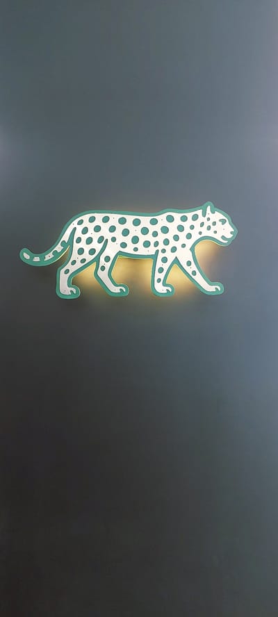 A neon-lit leopard cutout is affixed to a dark gray wall. The cutout is predominantly white with teal spots and a teal outline. The neon light, casting a warm yellow glow, emanates from behind the cutout, highlighting its edges and the wall immediately surrounding it. The leopard is depicted in a walking pose, with its body angled to the right. The background is a plain, textured dark gray wall that appears to be indoors. The lower portion of the image shows more of the dark gray wall, with no other objects or people visible. The scene is devoid of any identifiable urban or natural elements that would suggest a specific location within Lengkong Kulon, Indonesia.