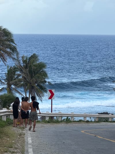 A group of four individuals stands by the roadside next to a palm tree in Burgos, Philippines. A white guardrail borders the asphalt road. Beyond the guardrail, there is a view of the ocean with waves. A red and white directional sign is next to the road.